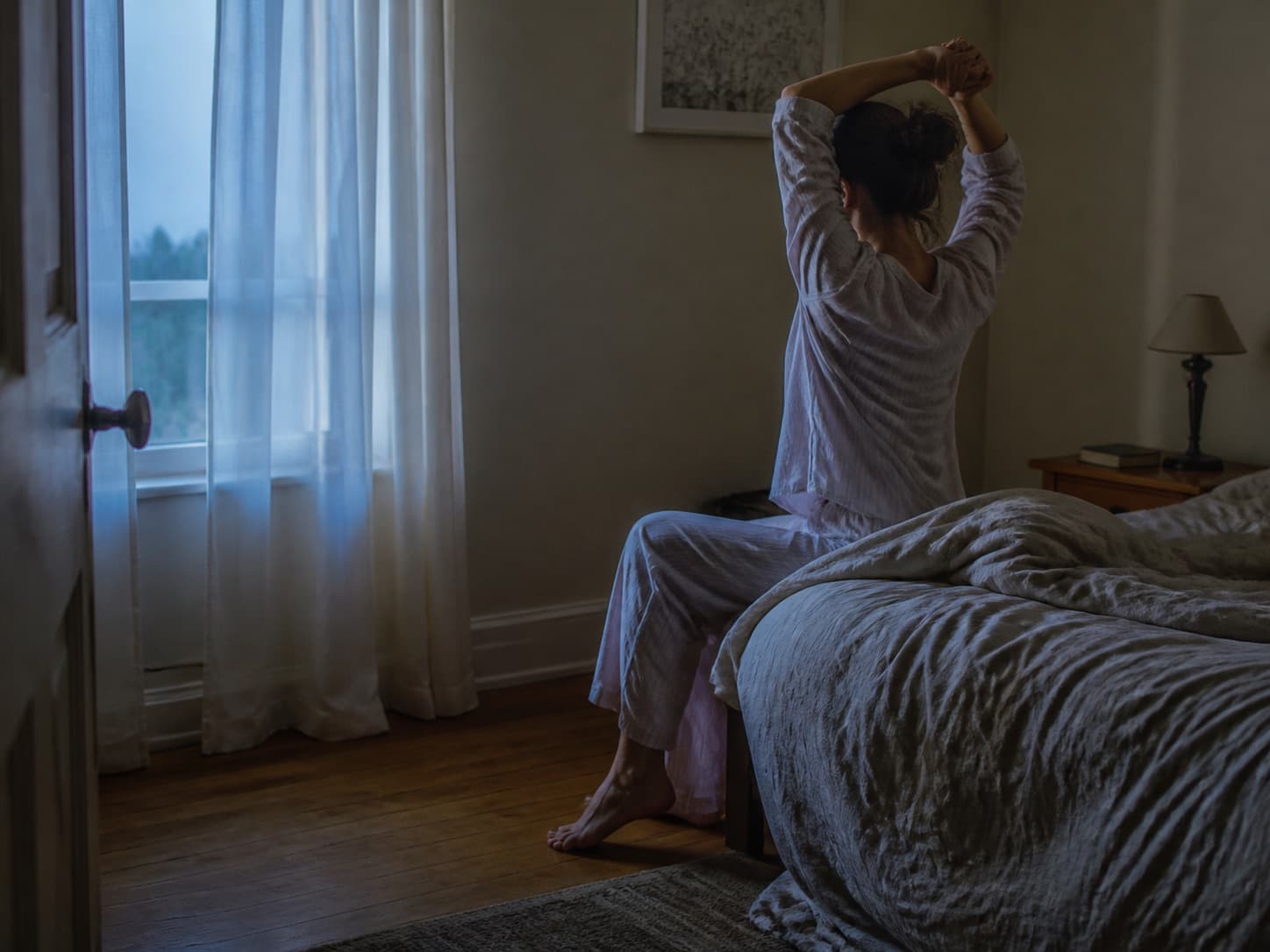 Woman stretching beside an unmade bed in gentle blue dawn light